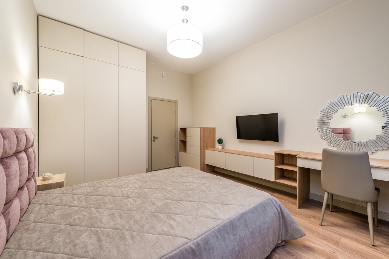 Interior of bedroom with comfortable bed under blanket placed near closet with TV set hanging on wall near mirror under lamp